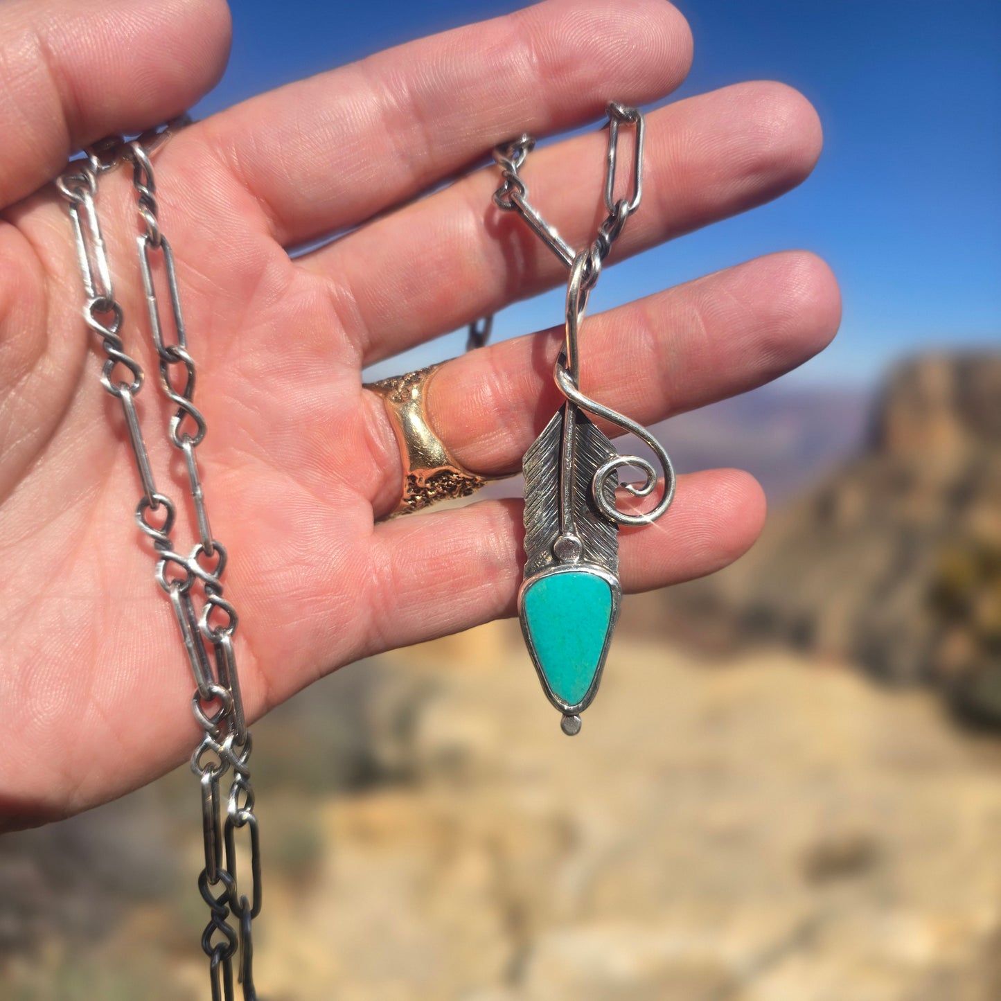 Hand stamped feather pendant in sterling silver featuring a turquoise accent, oxidized finish, and a large open bail, shown at approximately 2.5 inches long. Displayed on hand.