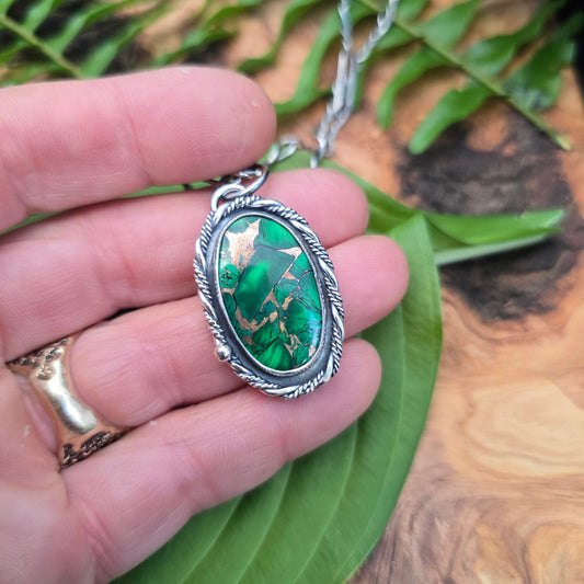 A silver-toned oval pendant necklace with a green marbled stone, held in a person's hand over a green leaf and wood surface.