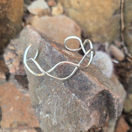 Sterling silver cuff bracelet with polished infinity symbol ends, shown on a natural surface.