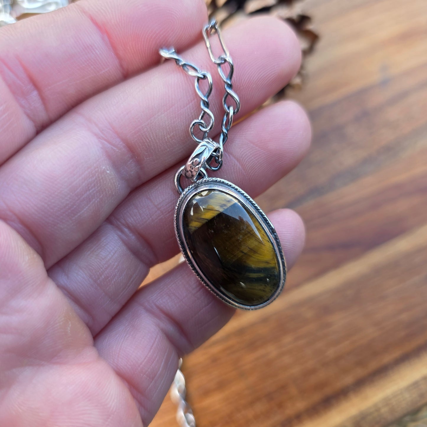 Tiger eye pendant featuring an oval stone with golden hues, wire perimeter detailing, and a flower-patterned bail, shown against a wooden surface on hand.