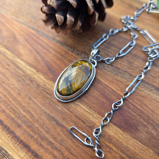 Tiger eye pendant with a flower-patterned bail and wire-wrapped perimeter, approximately 1.5 inches long, photographed flat on a wood surface.