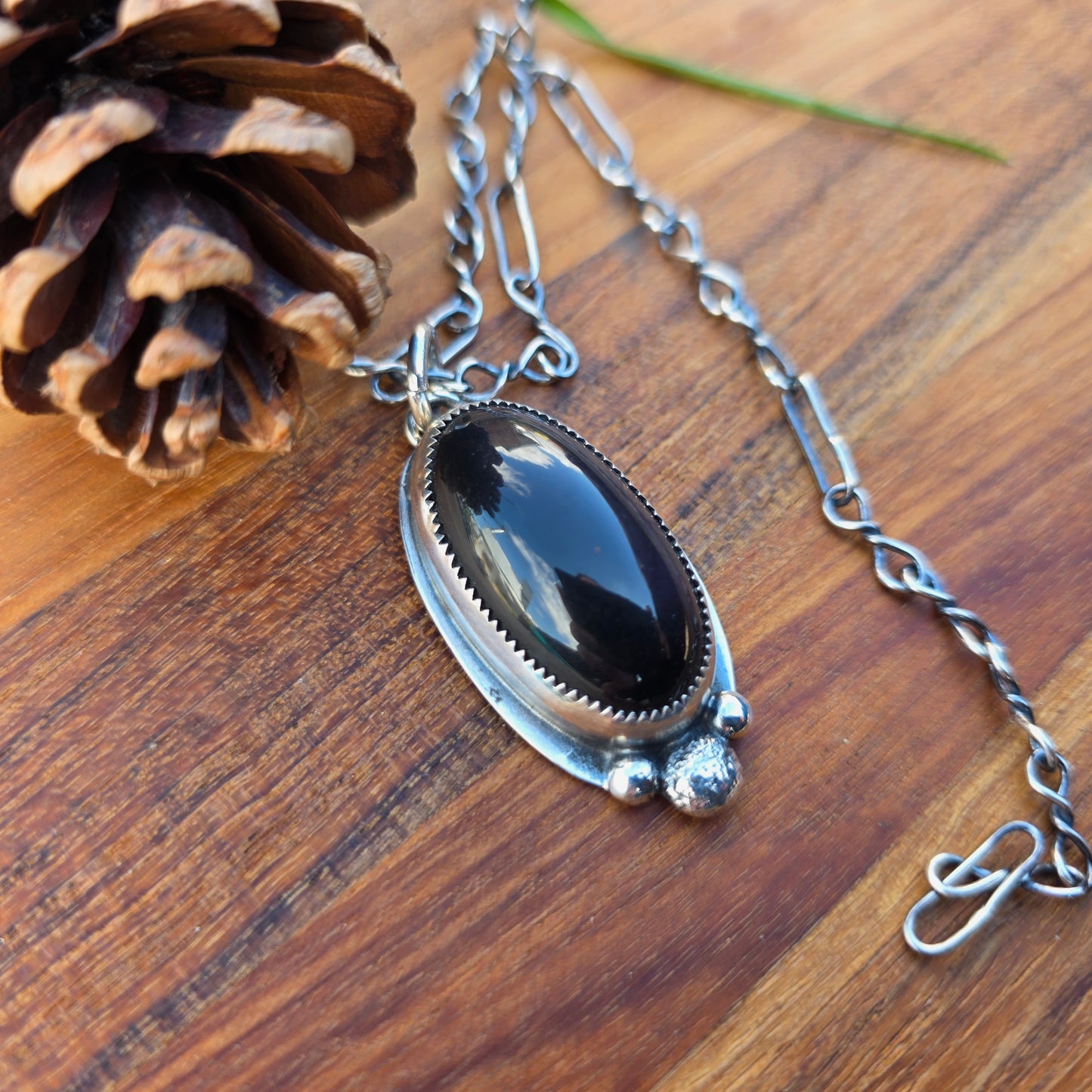 Three bead black obsidian pendant with an oval obsidian cabochon set in sterling silver, serrated bezel detail and backplate overhang visible, displayed on a wood surface, angled view.