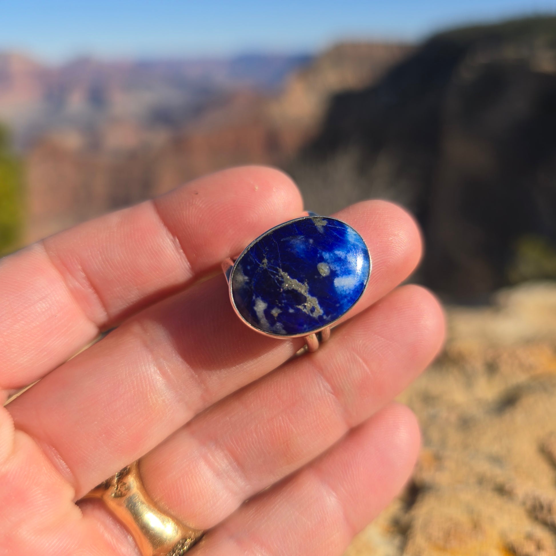 Oval blue sodalite stone set in a silver ring with a simple band design, displayed on a natural background on hand front view.