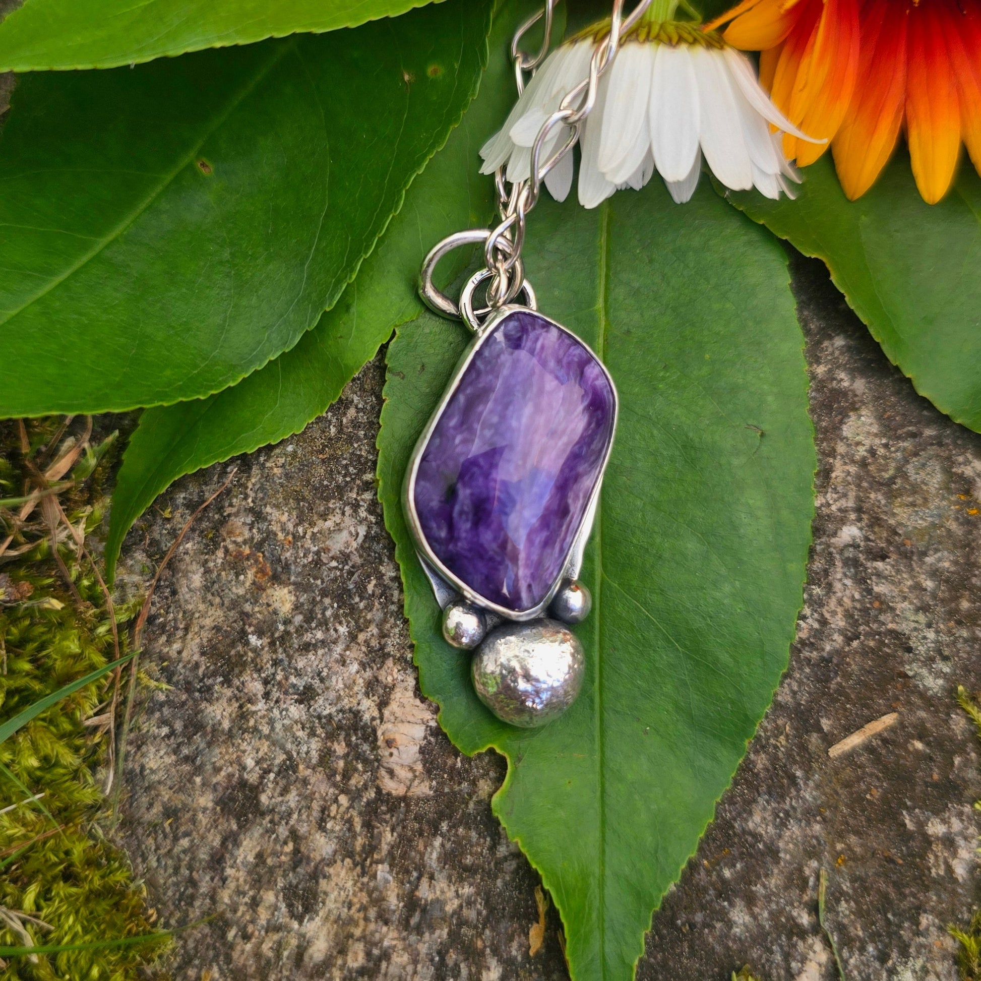 A silver pendant necklace with a polished purple amethyst stone and a small silver bead charm, displayed on green leaves and orange flowers over a rock outdoors.