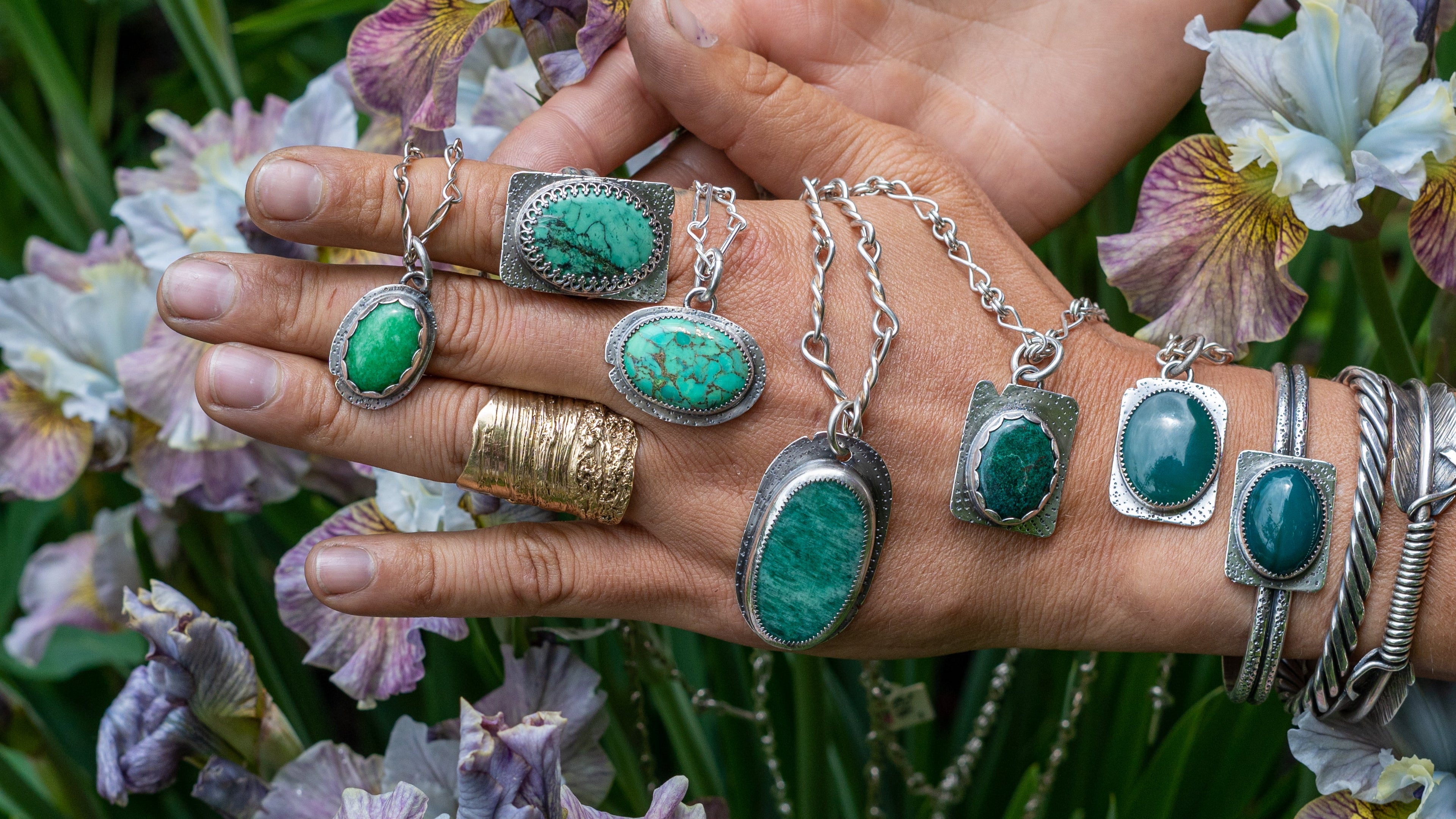 Collection of hand-forged jewelry displayed on hands with flowers in the background.