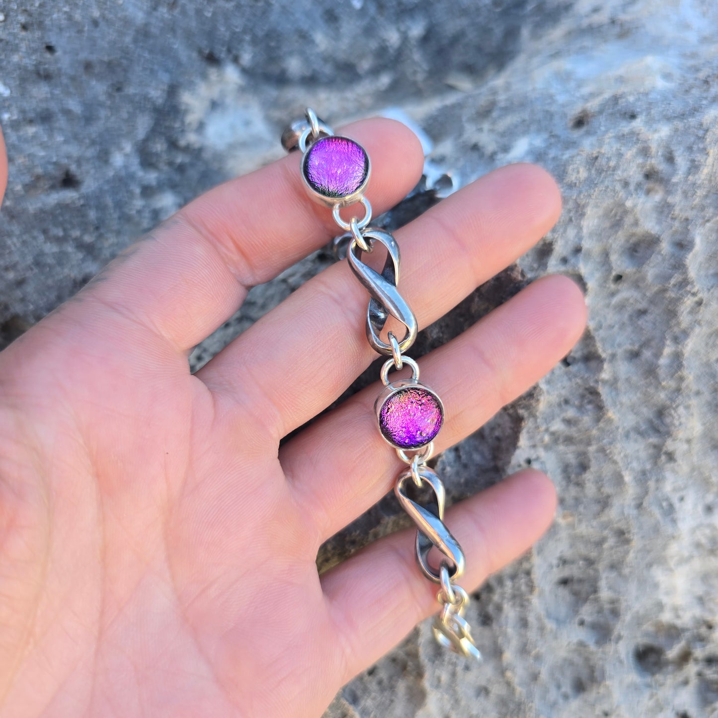 Sterling silver infinity bracelet with an oxidized finish and three pink dichroic stones, showing continuous infinity-shaped links shown on hand.