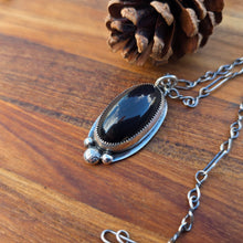 Load image into Gallery viewer, Black obsidian pendant with three round beads above an oval obsidian cabochon, set in sterling silver with a serrated bezel and visible backplate overhang, shown on a wooden surface, angled view
