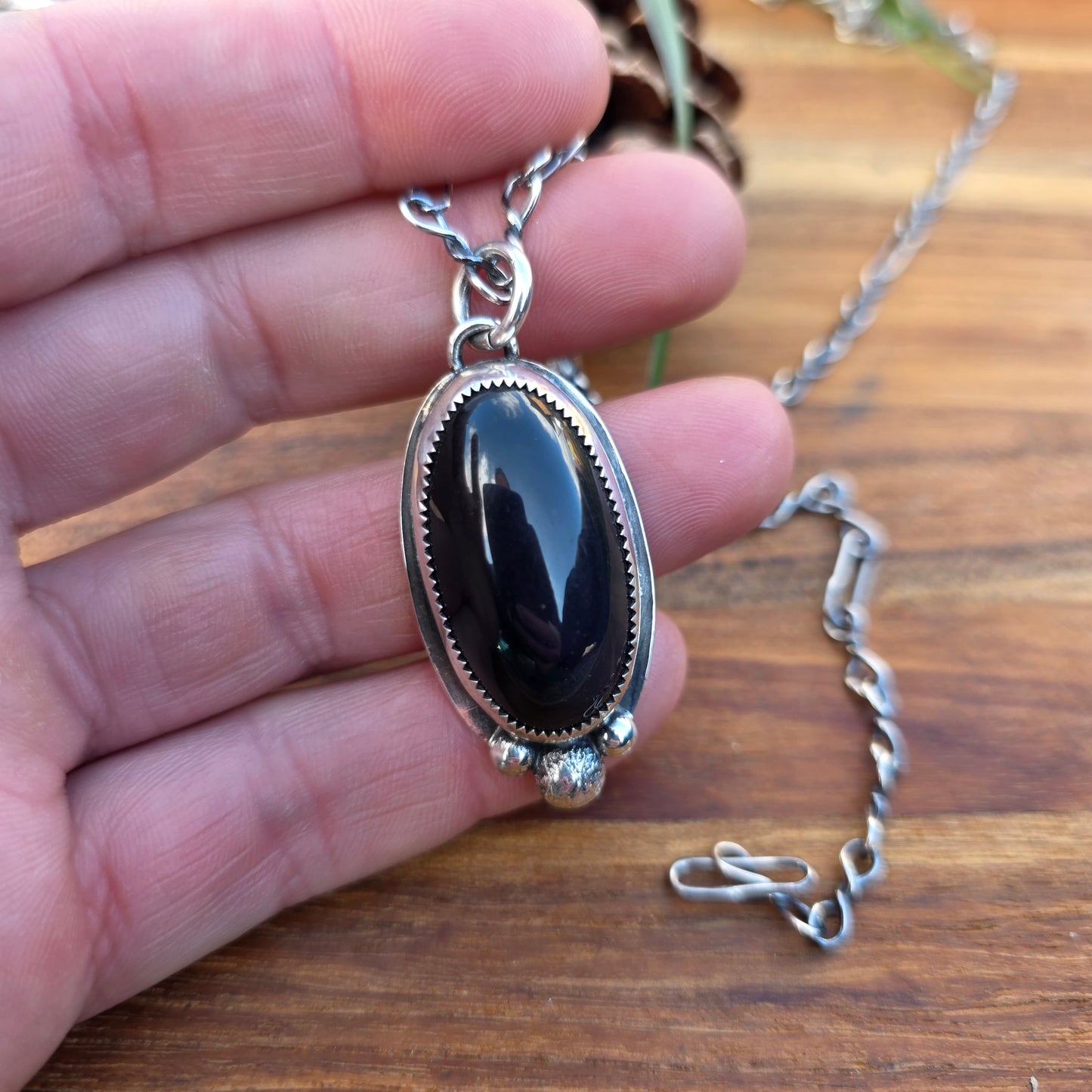 Black obsidian pendant with three stacked round beads and an oval cabochon centerpiece, sterling silver bezel with serrated edge and backplate visible, shown worn on hand.
