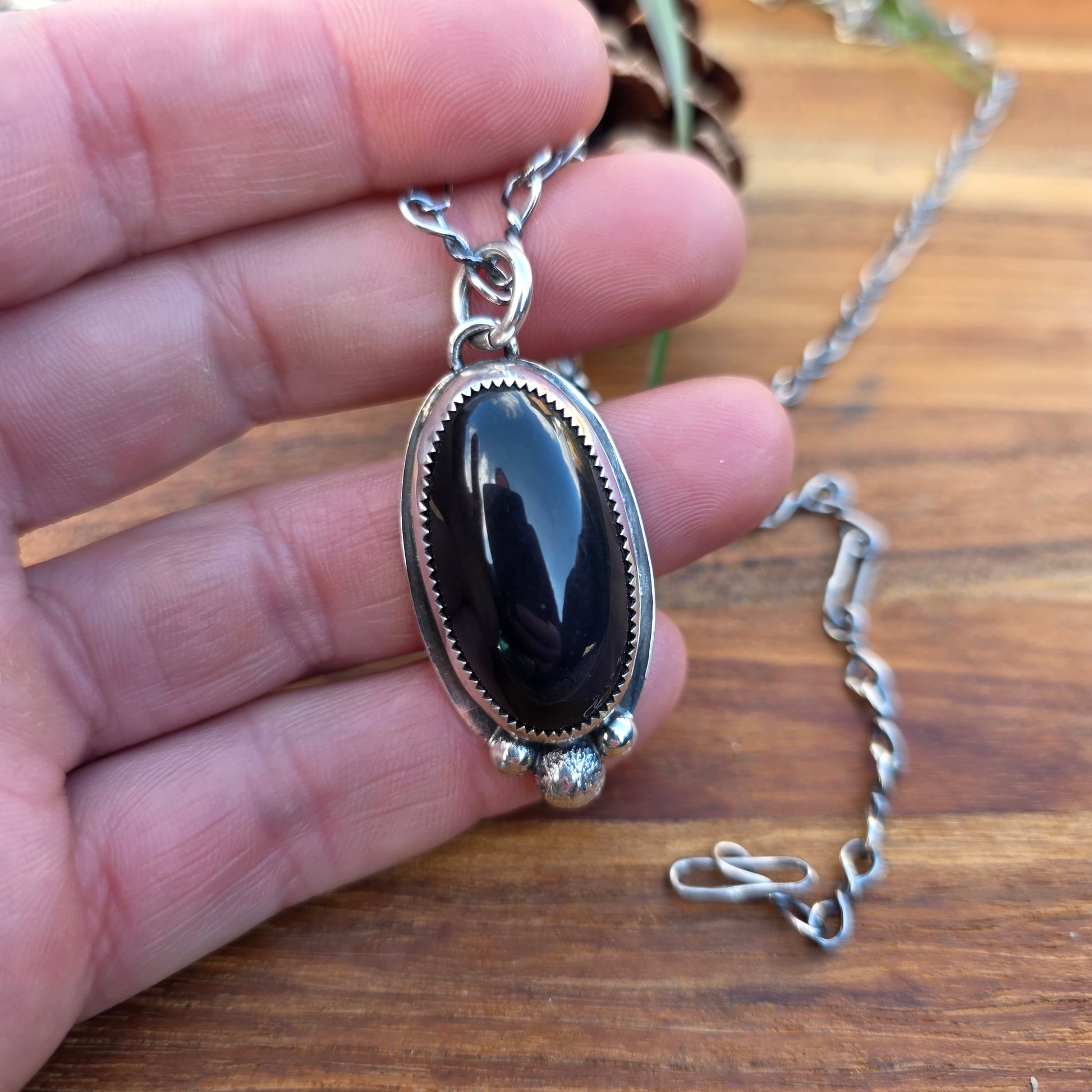 Black obsidian pendant with three stacked round beads and an oval cabochon centerpiece, sterling silver bezel with serrated edge and backplate visible, shown worn on hand.