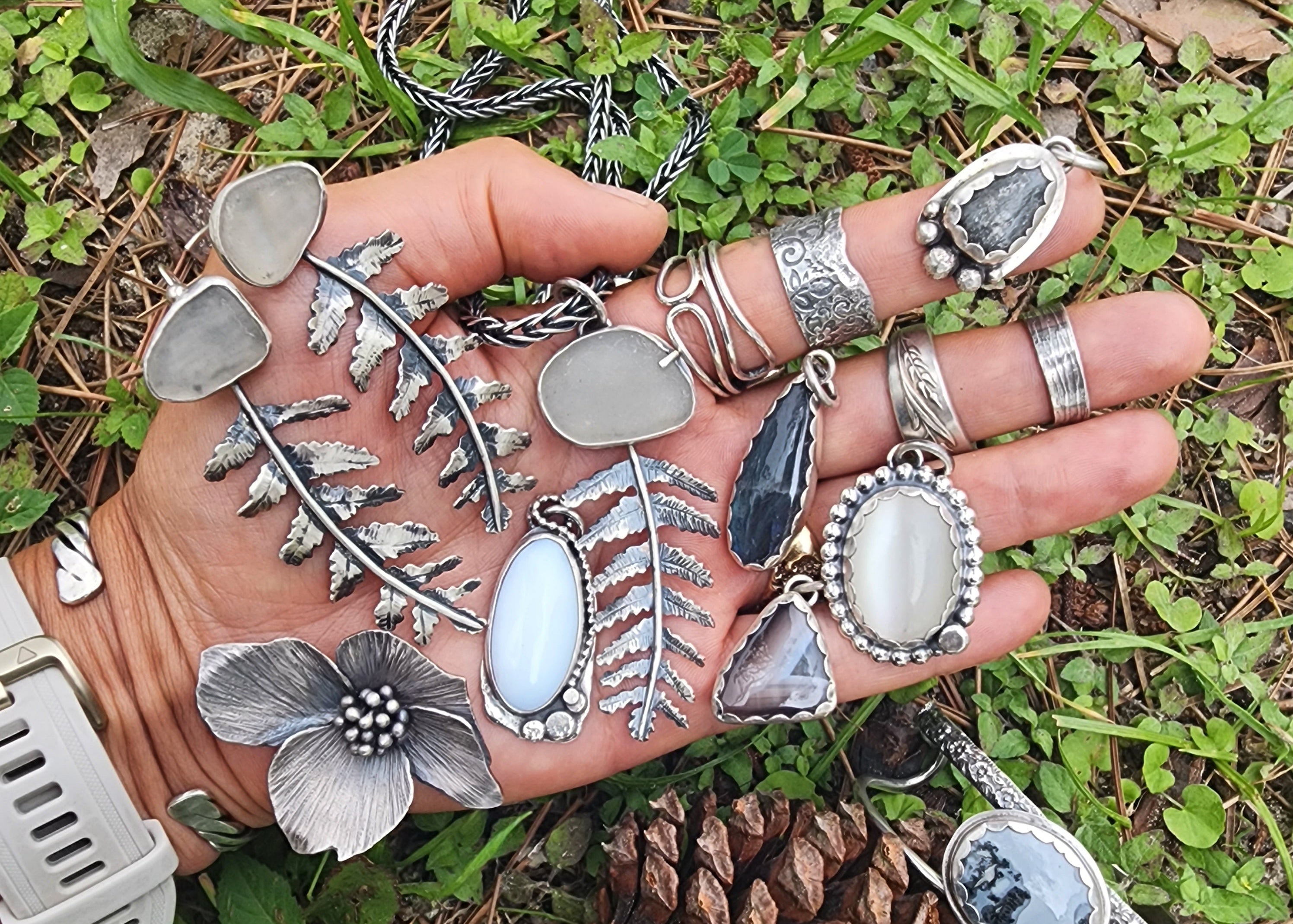 Collection of hand-forged jewelry including ferns, dogwoods, and white stones displayed on hand in natural setting