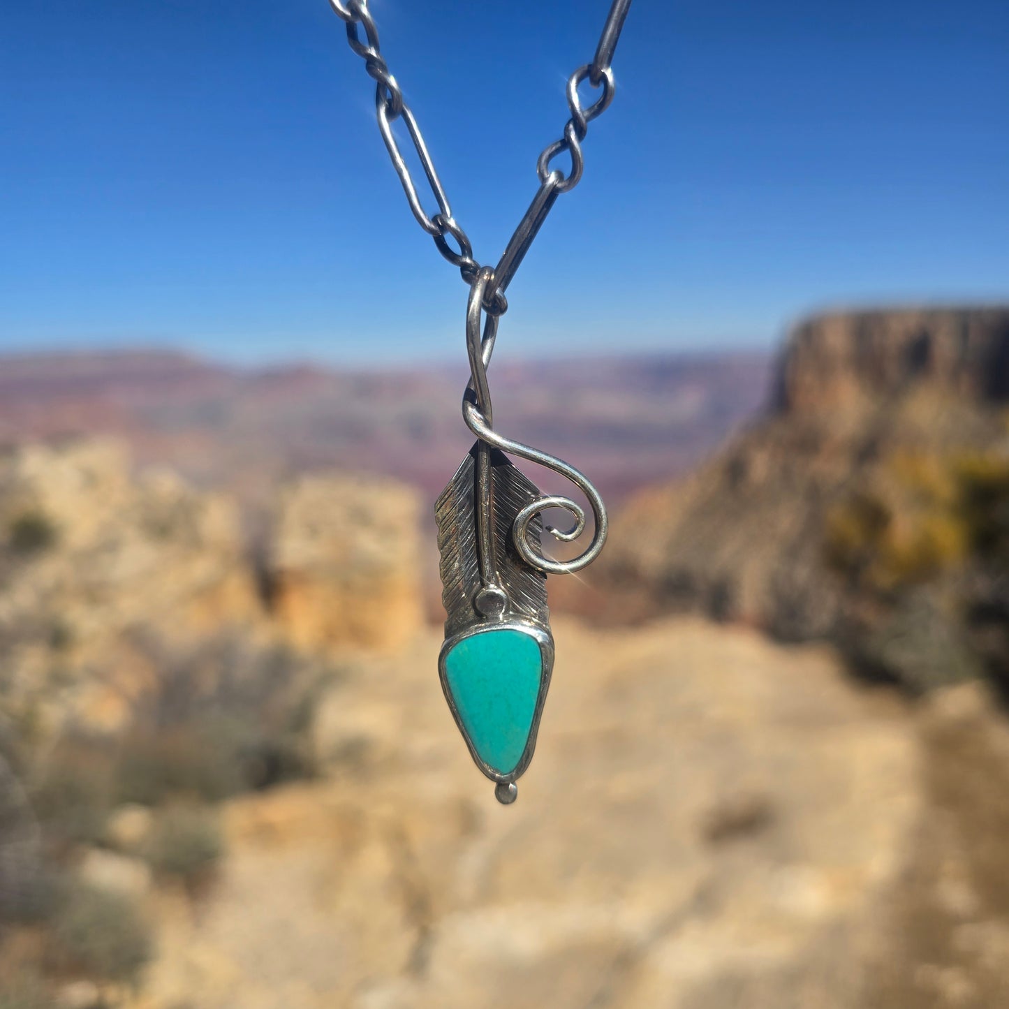 Sterling silver feather pendant with stamped texture and a turquoise accent stone, oxidized surface visible, measuring about 2.5 inches with a wide bail.