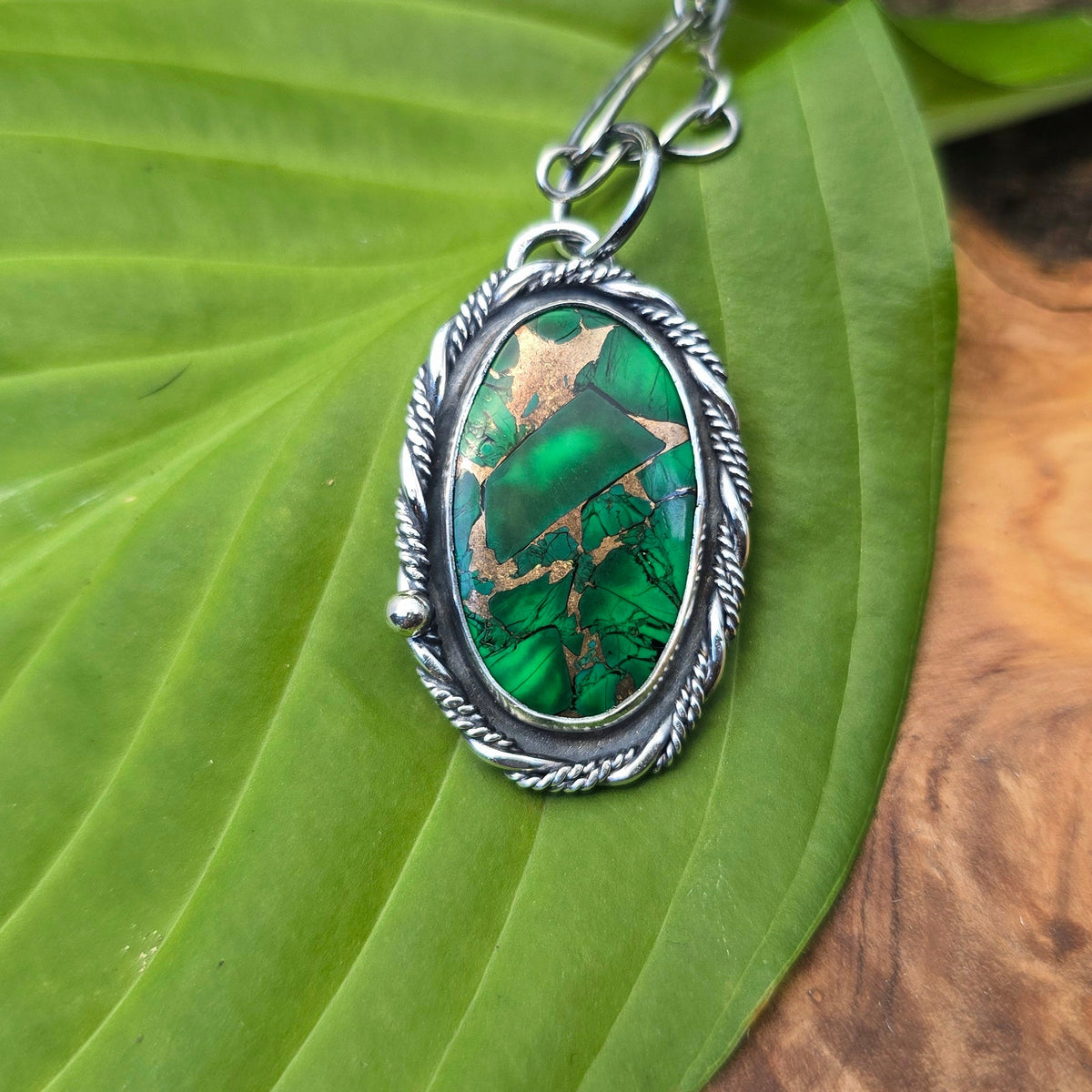 A silver-toned oval pendant necklace with a green marbled stone, resting on a large green leaf over a wood slice background.