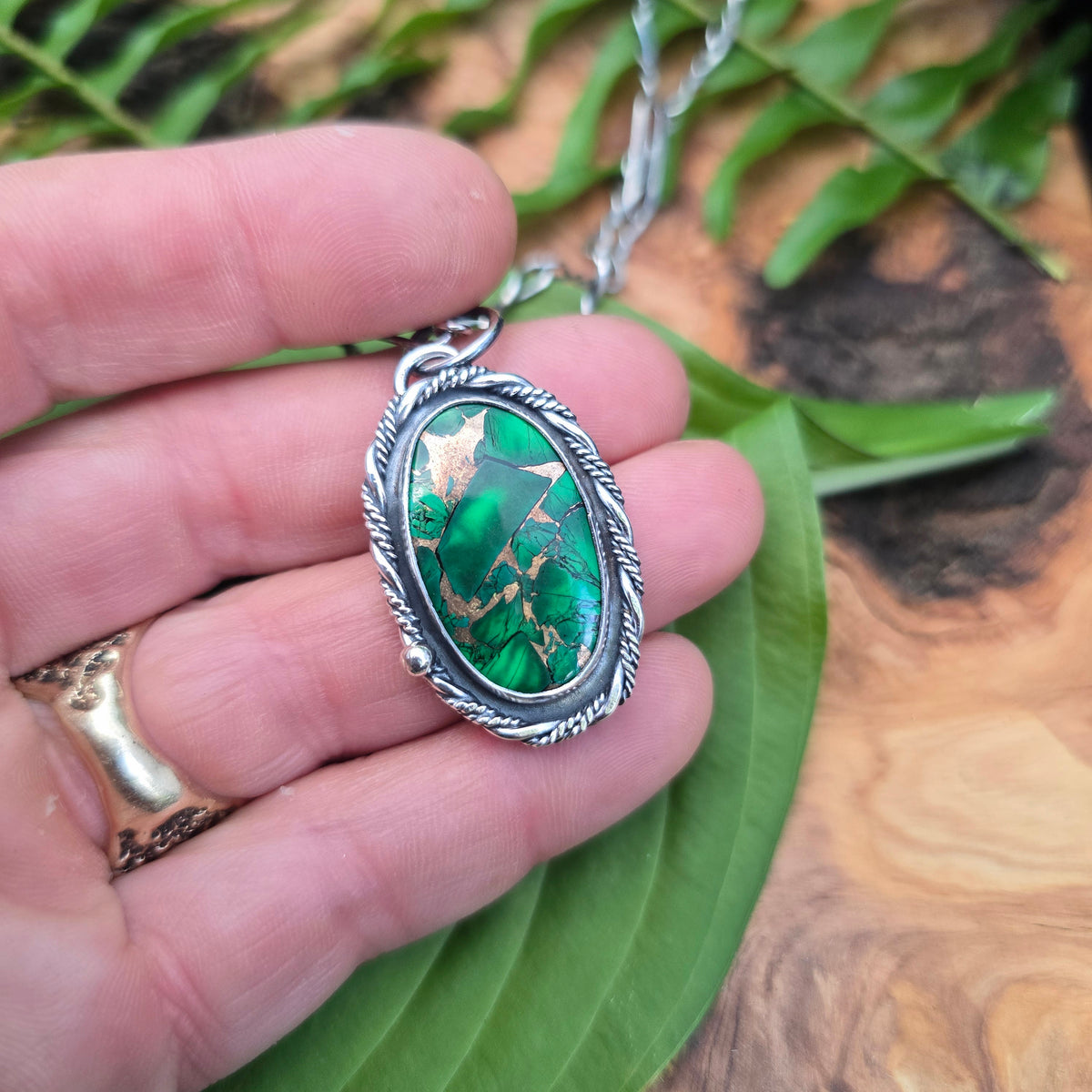 A silver-toned oval pendant necklace with a green marbled stone, held in a person's hand over a green leaf and wood surface.