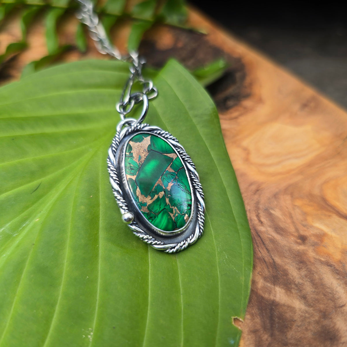 A silver-toned oval pendant necklace with a green malachite stone, resting on a large green leaf over a wood slice background.