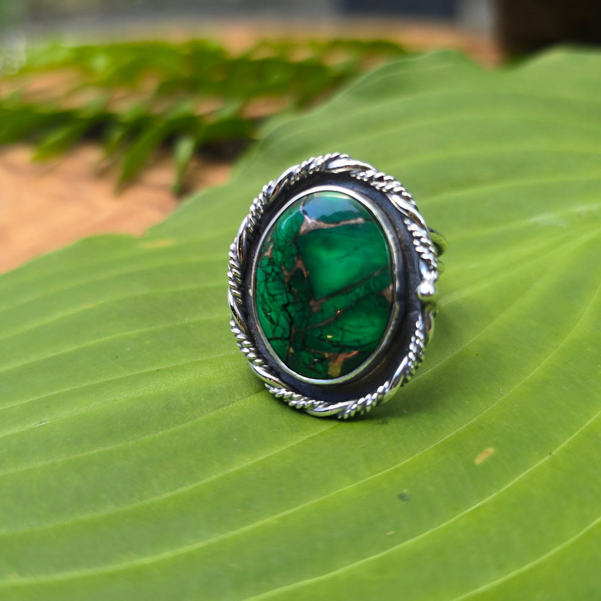 A silver ring with a round green malachite cabochon stone, resting on a large green leaf with a blurred natural background.
