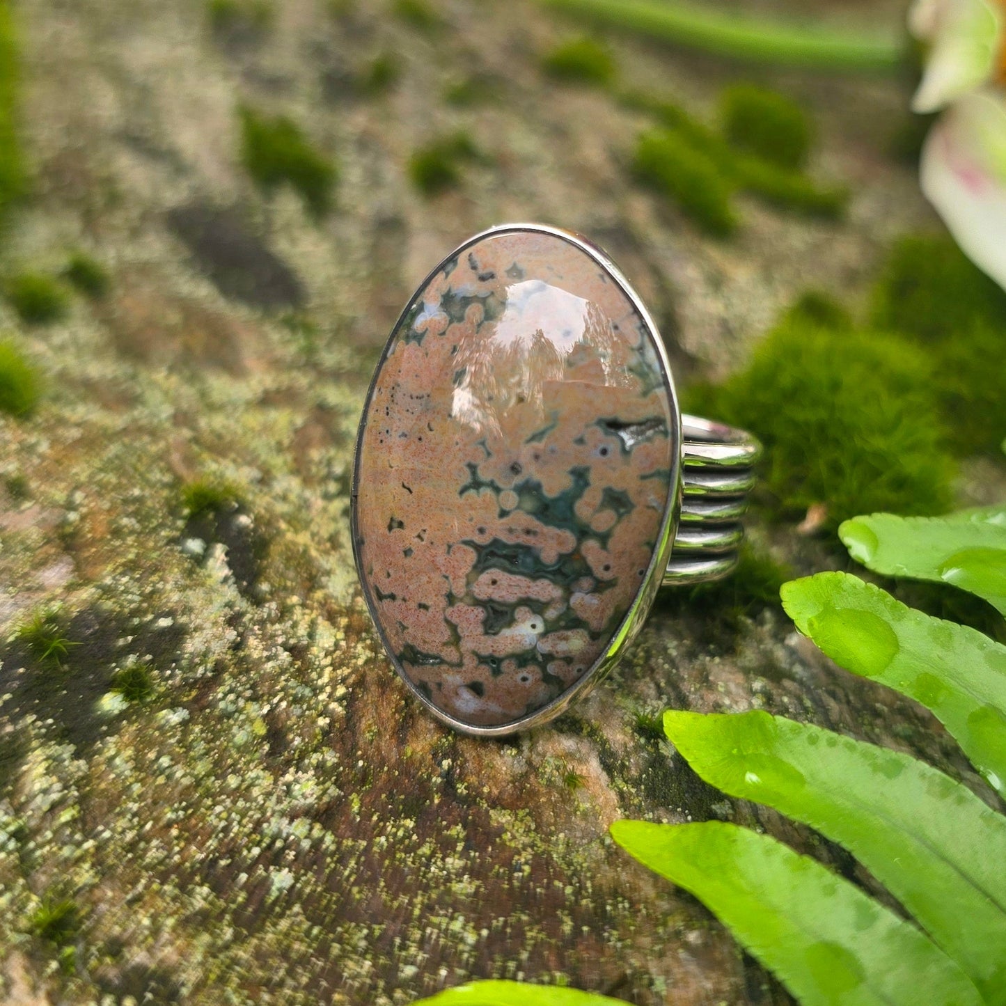 A silver ring with an oval polished stone featuring brown, beige, and black marbled patterns, displayed on a mossy rock with green ferns and blurred colorful flowers in the background.