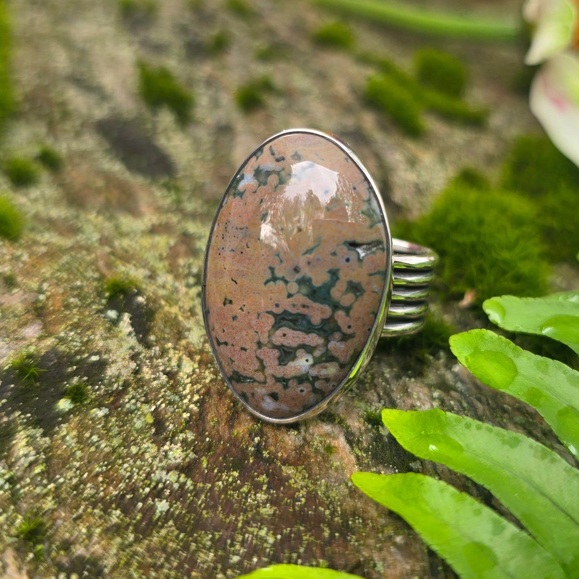 A silver ring with an oval polished stone featuring brown, beige, and black marbled patterns, displayed on a mossy rock with green ferns and blurred colorful flowers in the background.