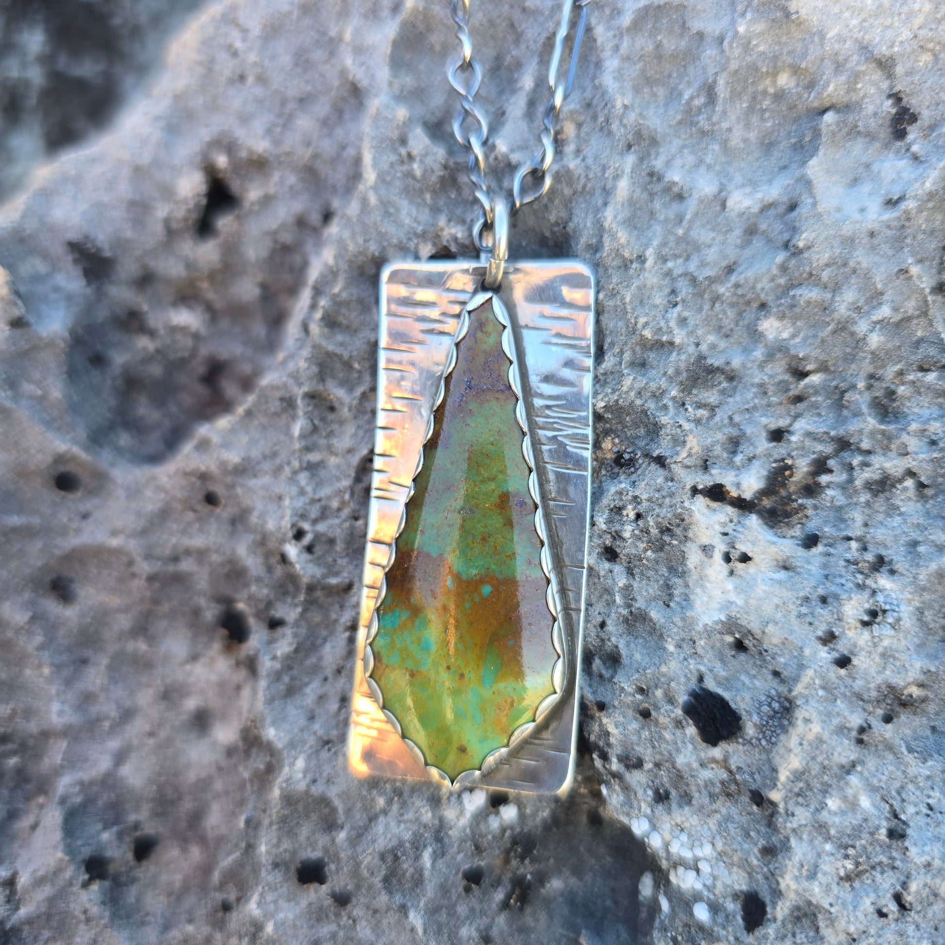 Green and brown Patagonia turquoise stone set in a silver pendant measuring about 2.25 inches, shown against a natural outdoor surface.