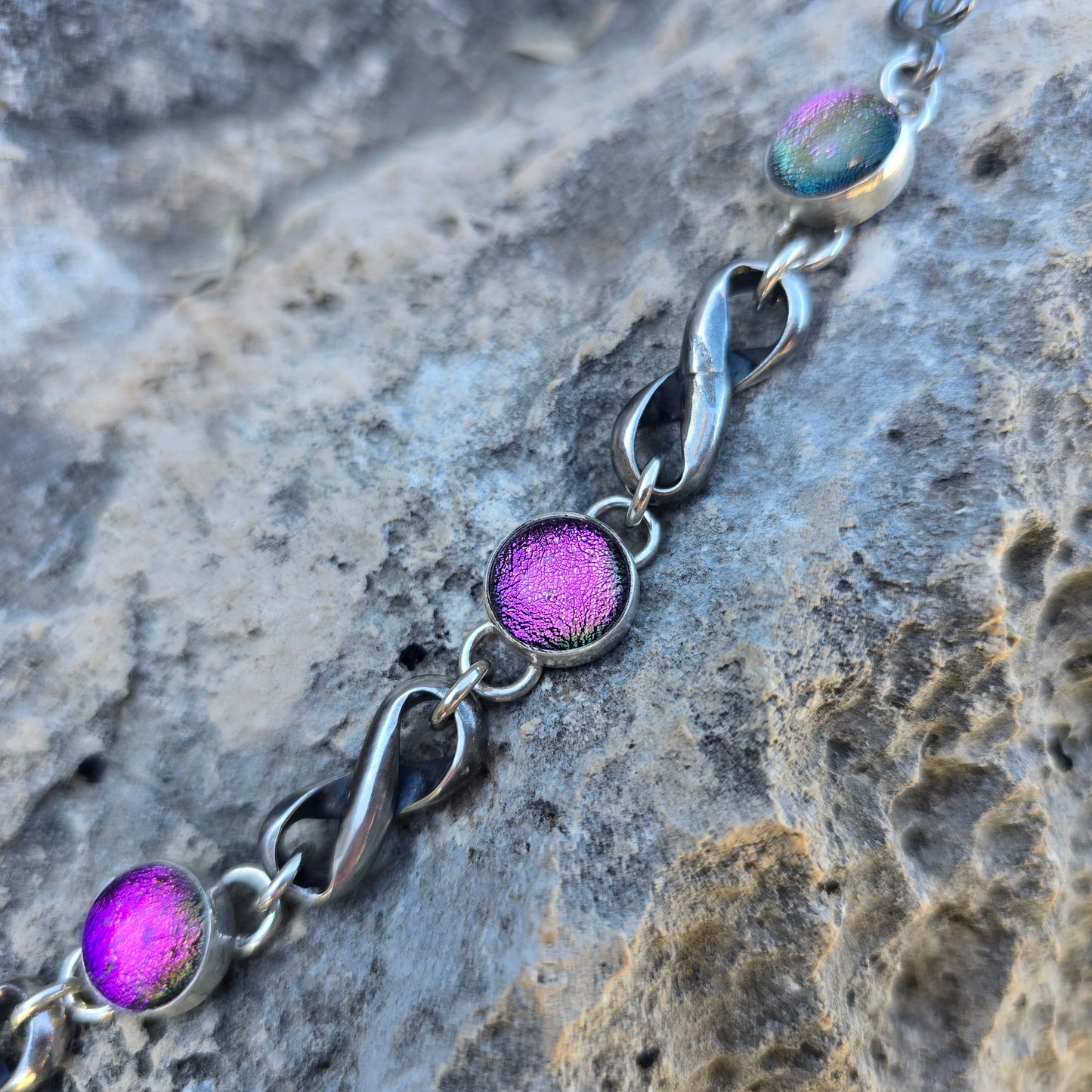 Oxidized sterling silver infinity bracelet featuring three pink dichroic stones, photographed flat with linked infinity elements visible close up view.