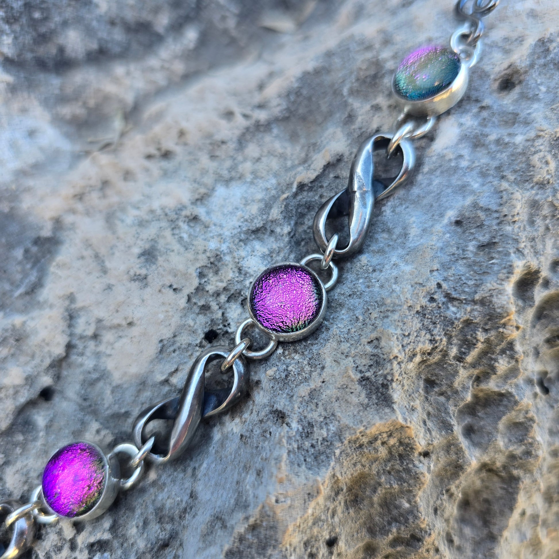 Oxidized sterling silver infinity bracelet featuring three pink dichroic stones, photographed flat with linked infinity elements visible close up view.
