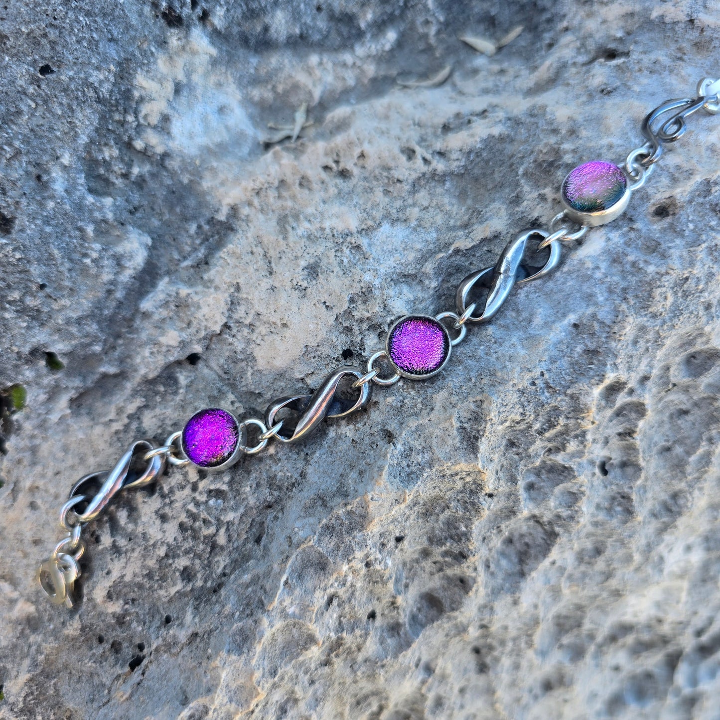 Infinity link bracelet with three pink dichroic glass stones set in oxidized sterling silver, shown with interconnected infinity-shaped links displayed on rock.