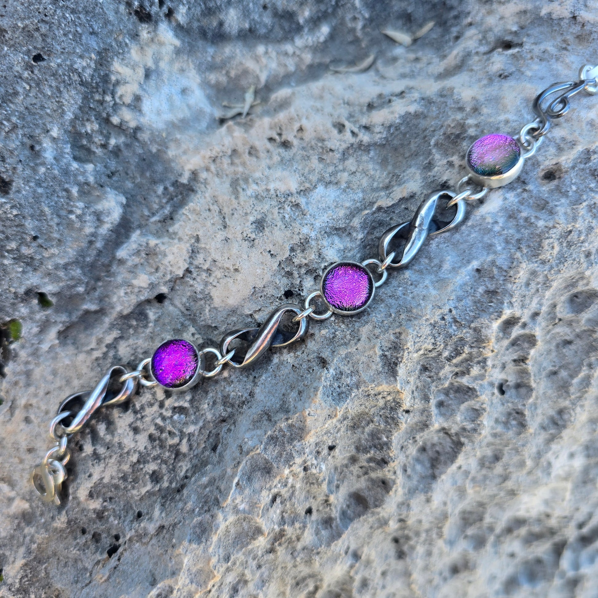 Infinity link bracelet with three pink dichroic glass stones set in oxidized sterling silver, shown with interconnected infinity-shaped links displayed on rock.