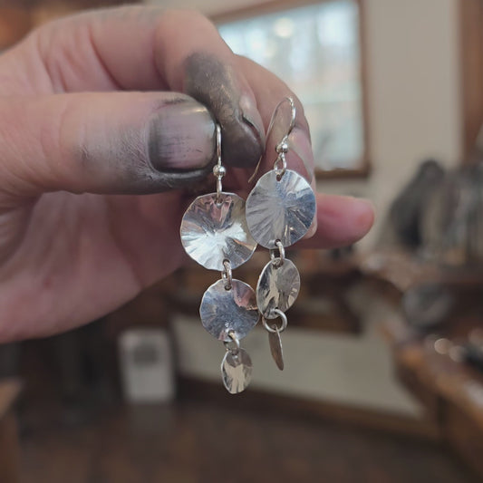 Pure silver hammered graduating disc earrings worn on hand, showing multiple round discs decreasing in size with a textured hammered surface and wire ear hooks.