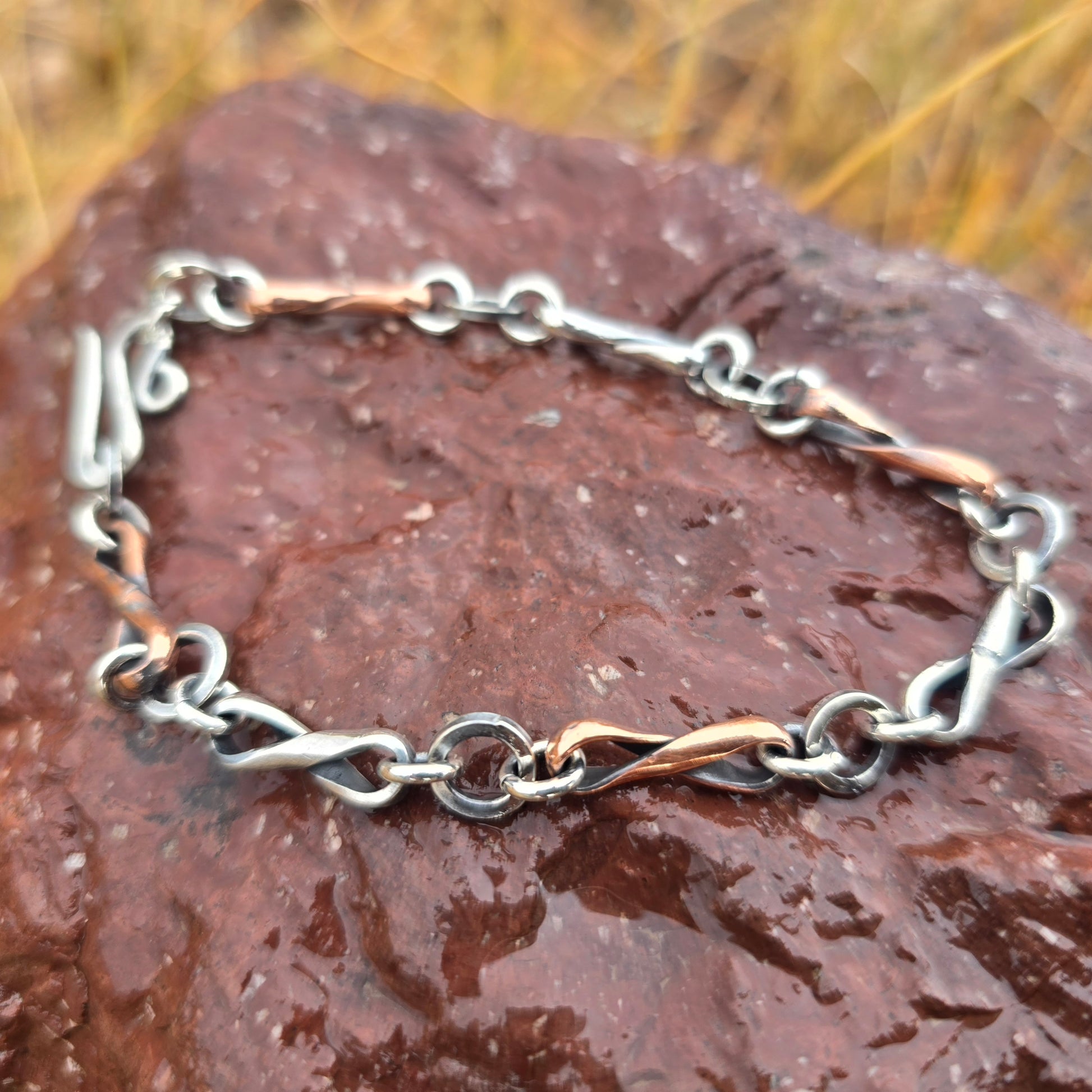 Alternating copper and sterling silver infinity link bracelet made from half round wire, oxidized finish, shown with a hand-forged clasp in a natural outdoor setting.