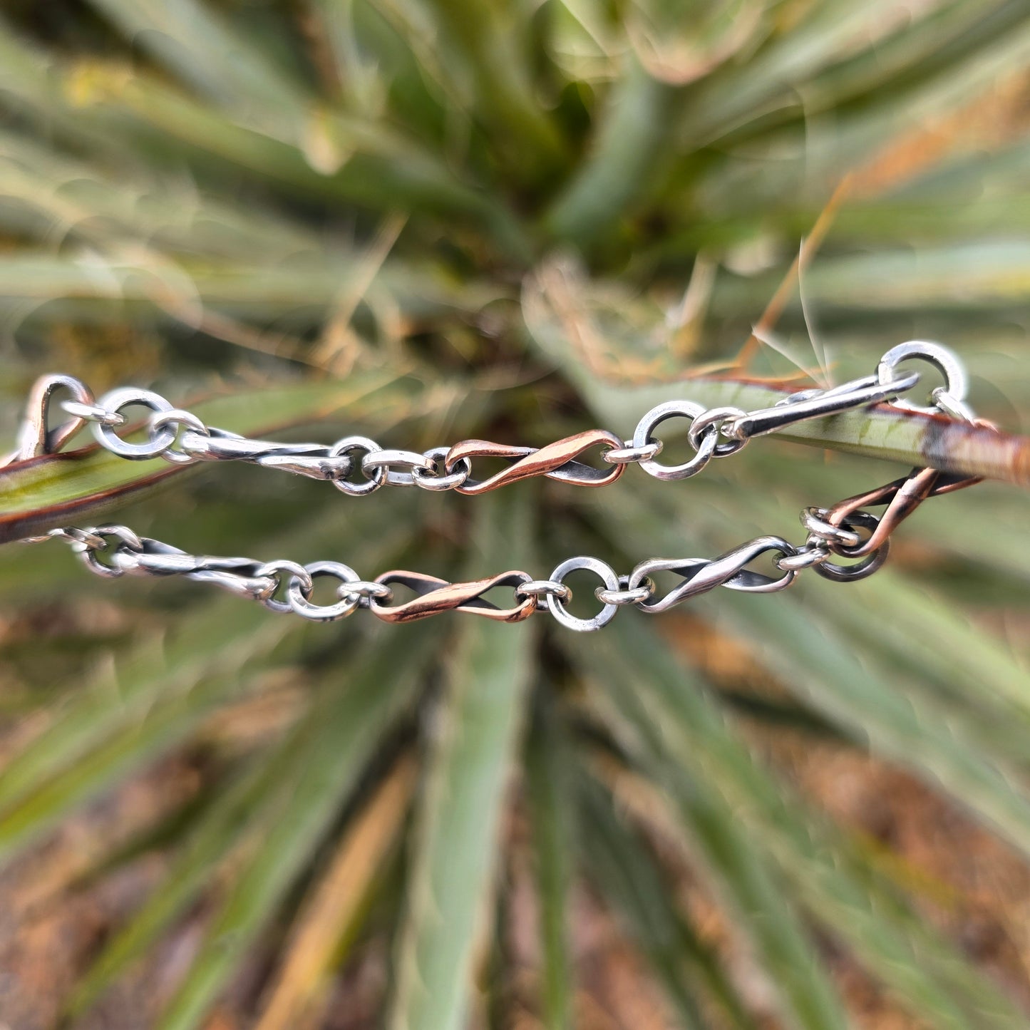Alternating copper and sterling silver infinity link bracelet constructed from half round wire, oxidized surface visible, with a hand-forged clasp shown outdoors.