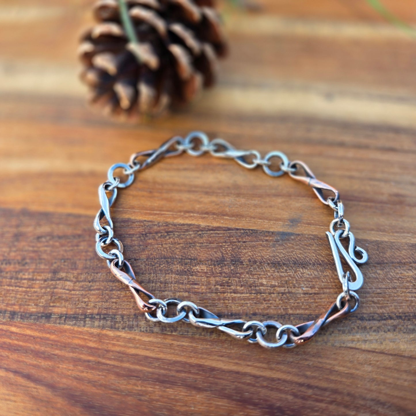 Alternating copper and sterling silver infinity link bracelet made from half round wire, oxidized finish, shown with a hand-forged clasp in a natural outdoor setting top view.