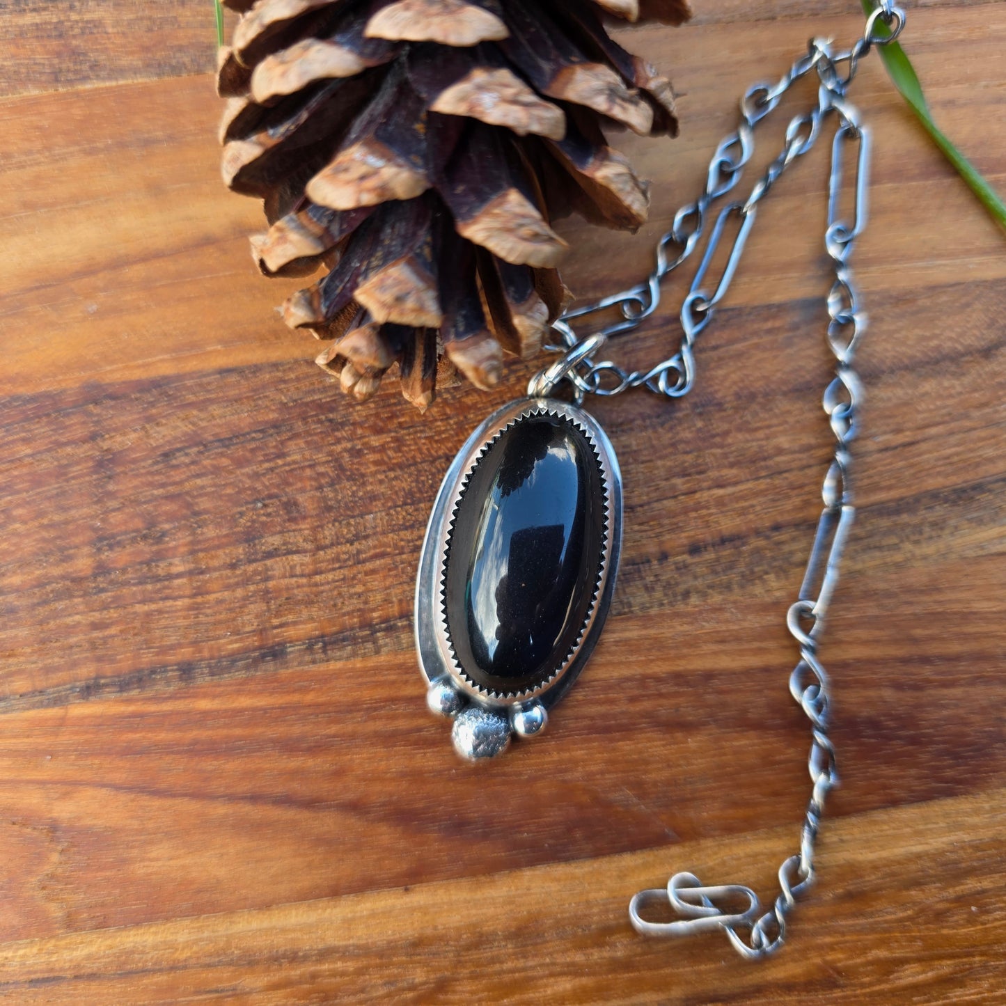 Black obsidian three bead pendant with a thick oval cabochon, sterling silver serrated bezel and flat backplate visible, shown on wood background, top view.