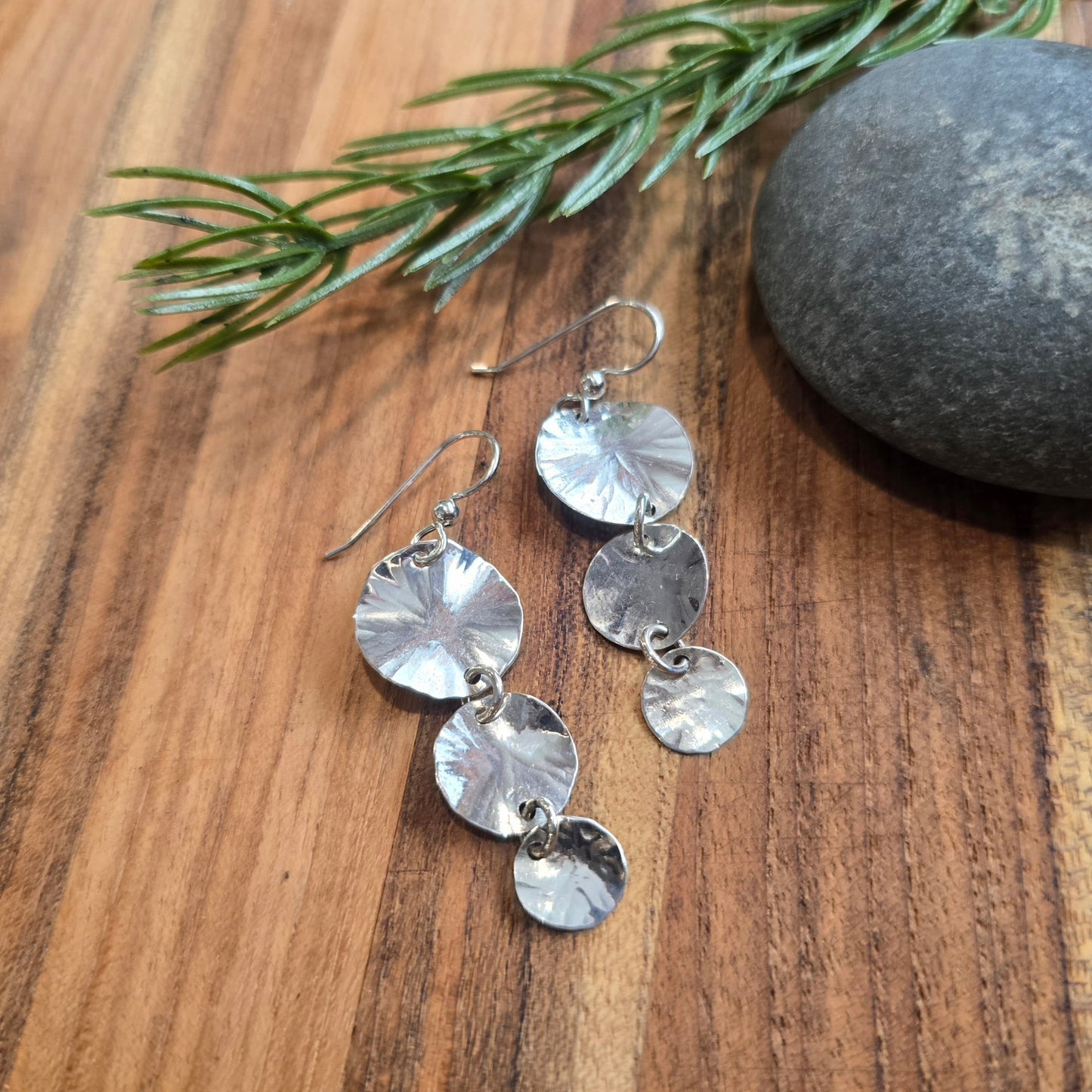 Pure silver hammered graduating disc earrings displayed flat on a board in top view, showing stacked round discs with a hammered texture and wire ear hooks.