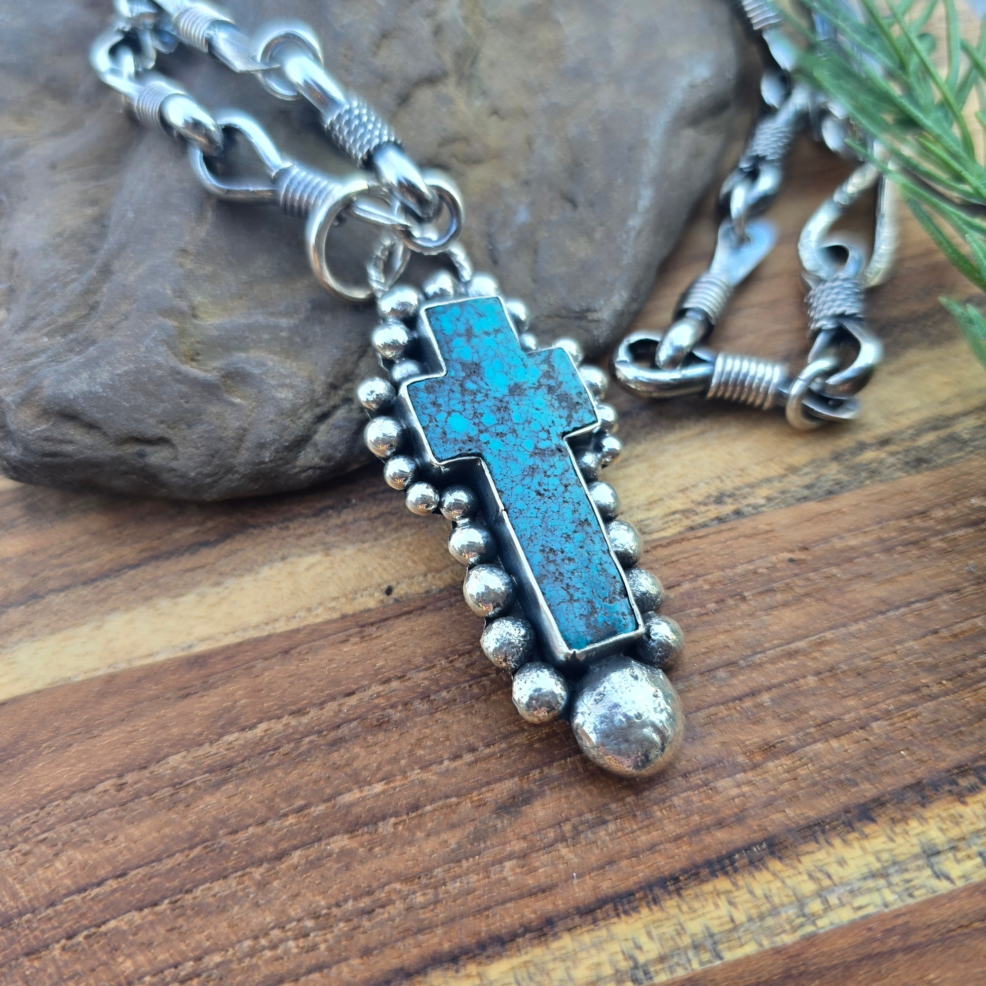 Angled view of a sterling silver cross pendant with beaded detailing, set with turquoise and finished with an oxidized surface.