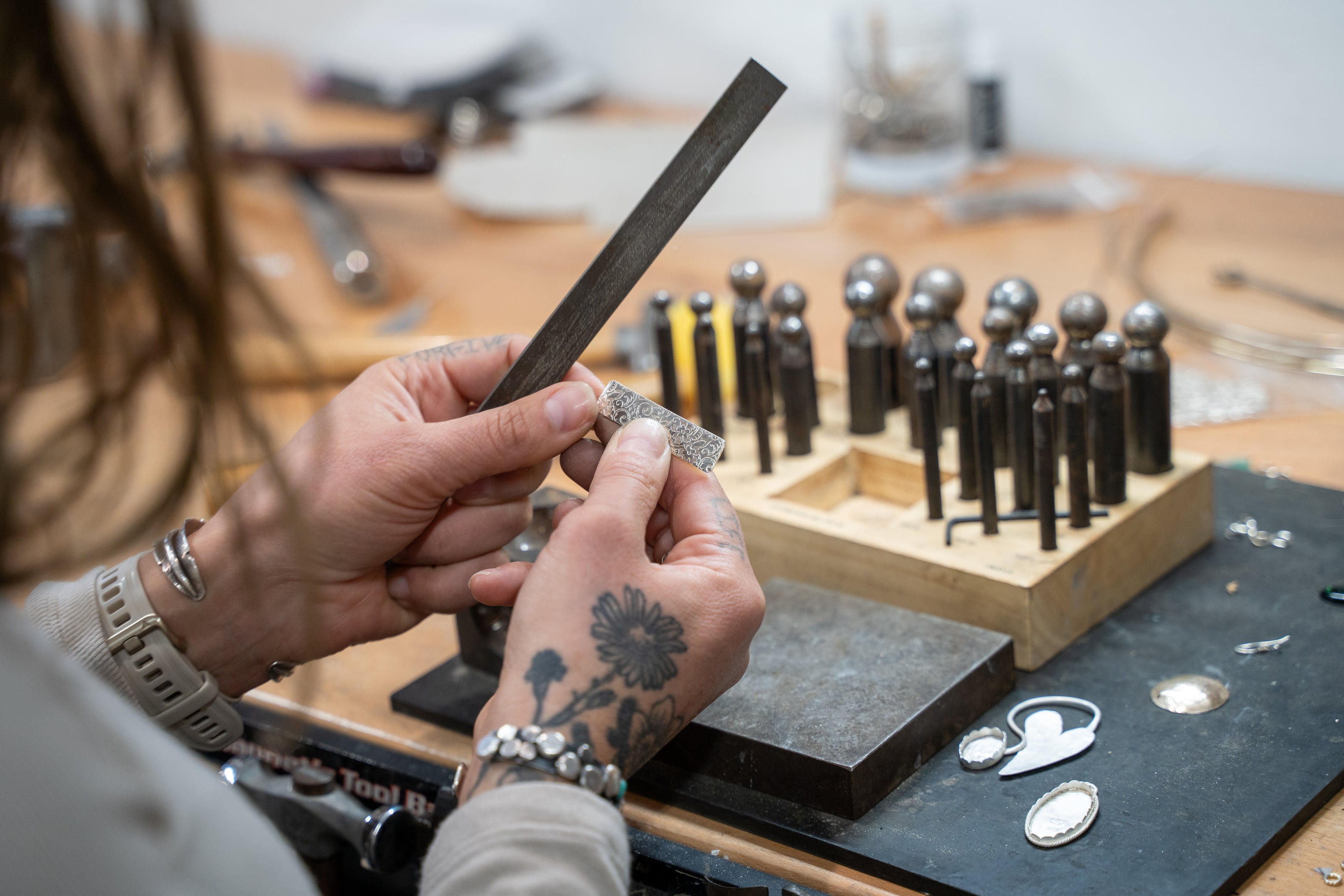 Adirondack Silversmith forging sterling silver jewelry using traditional file with daping block and anvil in background on workbench.
