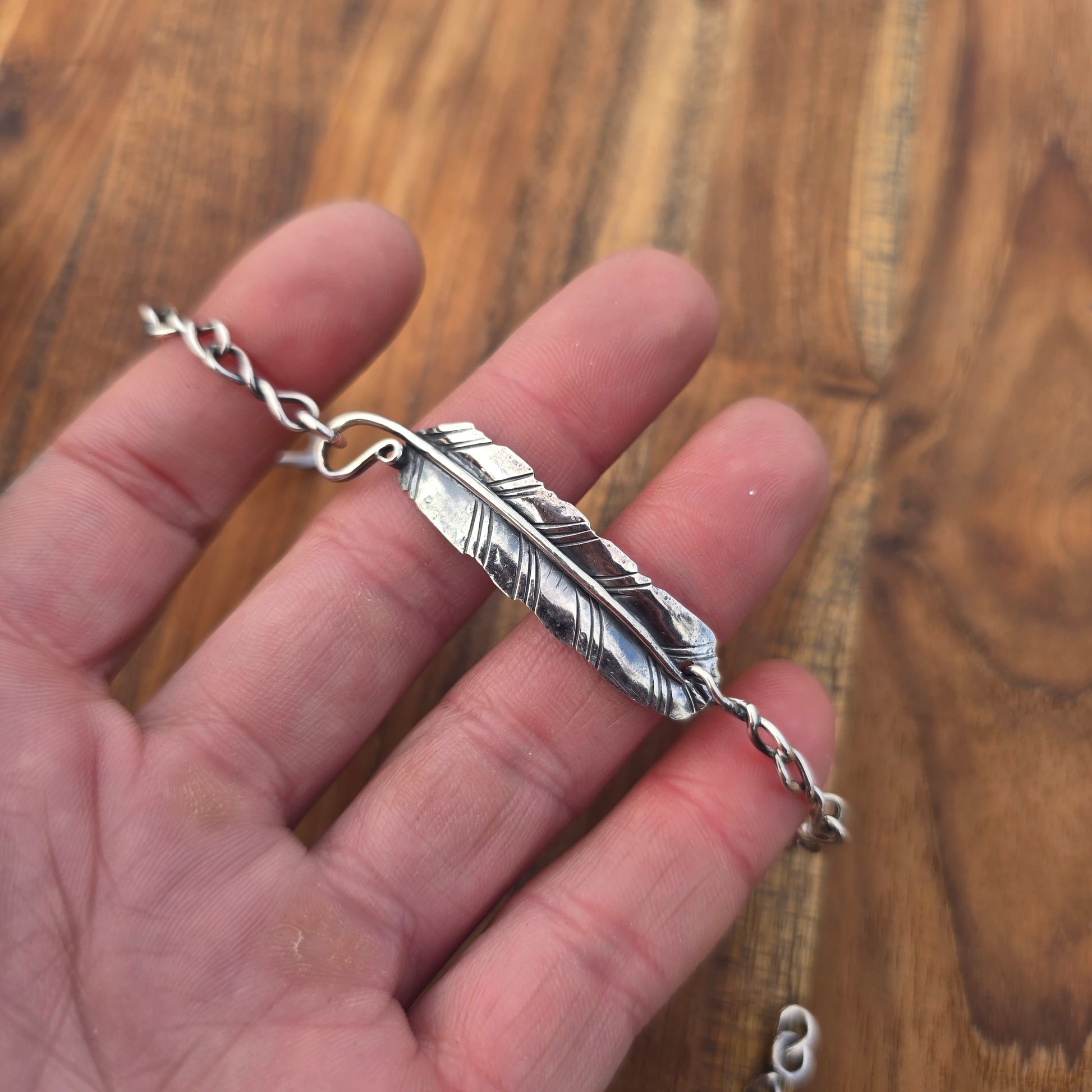Silver feather-shaped pendant on a chain held in a hand against a wooden background