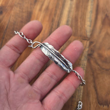 Load image into Gallery viewer, Silver feather-shaped pendant on a chain held in a hand against a wooden background
