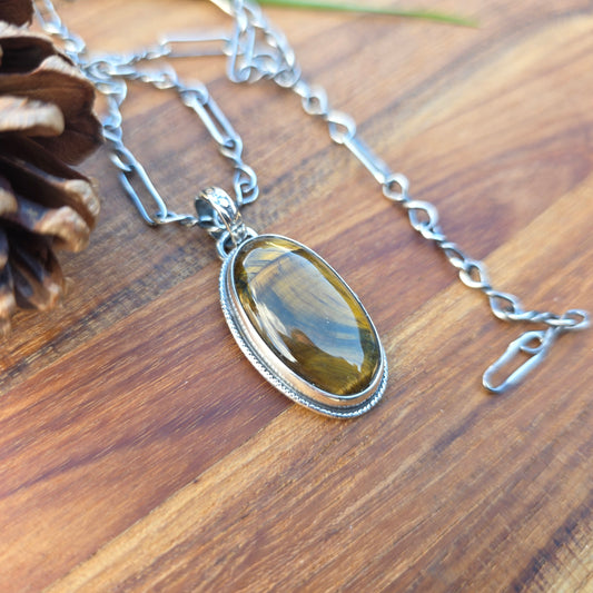 Oval tiger eye pendant with golden brown banding, decorative wire perimeter, and floral-patterned bail, shown on a wooden background.