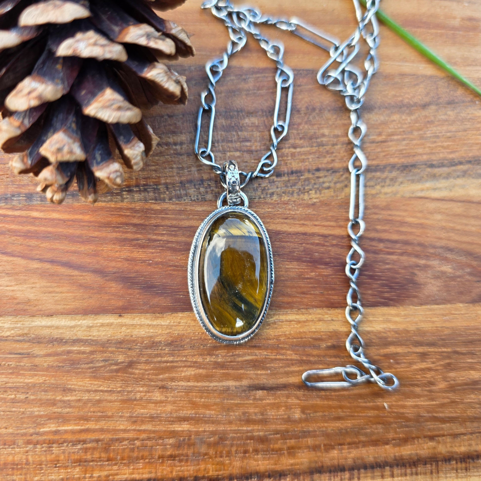 Tiger eye pendant measuring approximately 1.5 inches, featuring a flower-patterned bail and decorative wire perimeter, shown on a wooden background with golden brown stone banding visible.