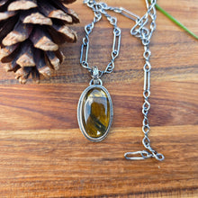 Load image into Gallery viewer, Tiger eye pendant measuring approximately 1.5 inches, featuring a flower-patterned bail and decorative wire perimeter, shown on a wooden background with golden brown stone banding visible.
