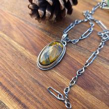 Load image into Gallery viewer, Tiger eye pendant with a flower-patterned bail and wire-wrapped perimeter, approximately 1.5 inches long, photographed flat on a wood surface.
