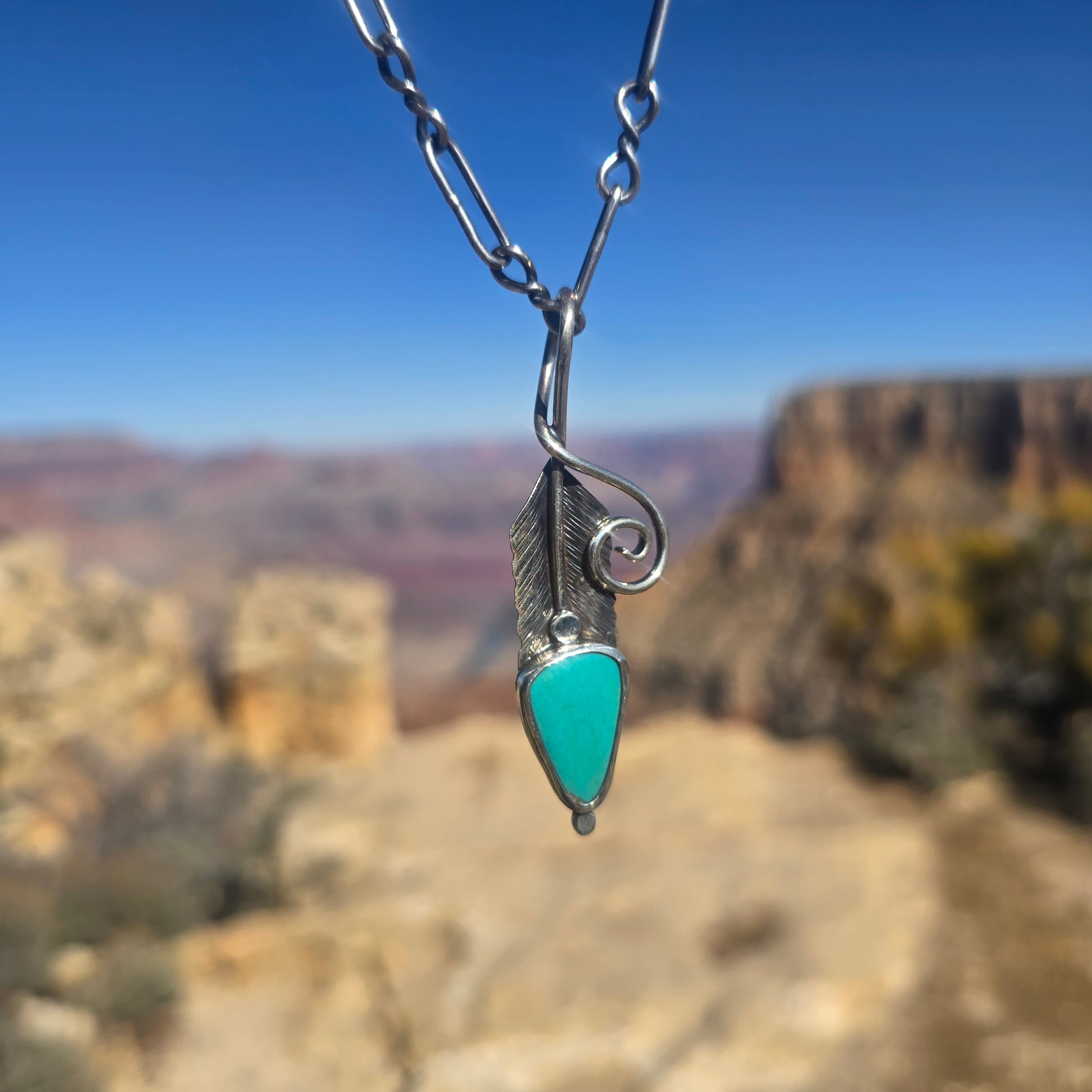 Feather-shaped sterling silver pendant with hand stamped detailing, turquoise accent, oxidized finish, and a large bail, approximately 2.5 inches in length displayed in front on natural background.