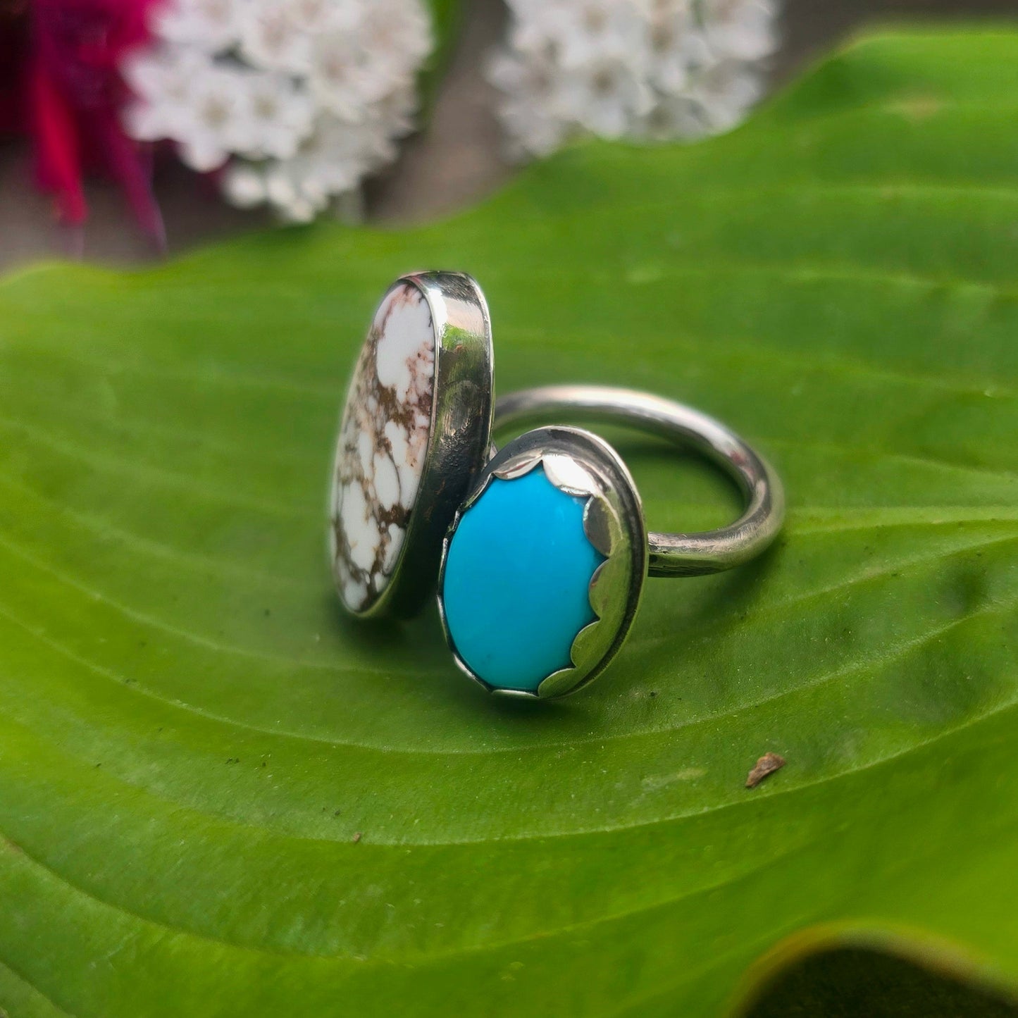 A sterling silver ring with two oval turquoise stone settings, one featuring a white and brown marbled pattern and the other a solid bright turquoise blue, displayed on a green leaf with blurred flowers in the background.